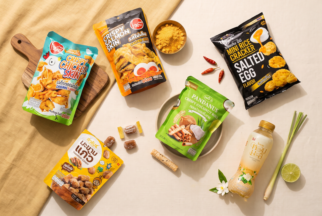 Six PLB Thai snack products on cream and mustard linen with brass bowl of salted-egg powder, Thai chillies, lemongrass and lime.